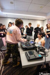A person demonstrating DJ equipment to a group in a room with "Pioneer DJ" gear visible on the table. The group appears attentive, standing and observing the setup.