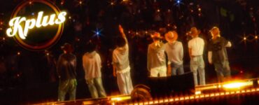 "Seven individuals stand on a stage facing an audience, with one raising a hand. They are wearing casual clothes and hats. Bright lights create a starry effect. The word 'Kplus' is visible in the upper left corner."