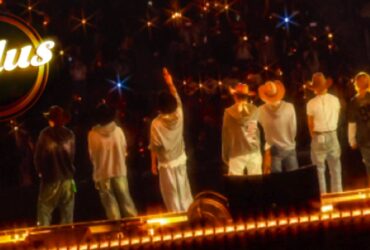 "Seven individuals stand on a stage facing an audience, with one raising a hand. They are wearing casual clothes and hats. Bright lights create a starry effect. The word 'Kplus' is visible in the upper left corner."