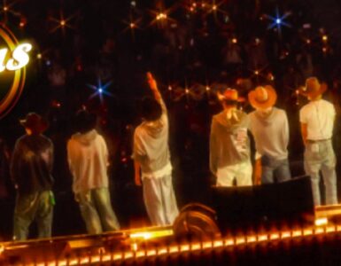 "Seven individuals stand on a stage facing an audience, with one raising a hand. They are wearing casual clothes and hats. Bright lights create a starry effect. The word 'Kplus' is visible in the upper left corner."