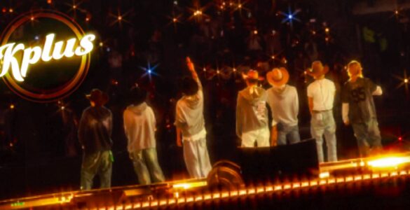 "Seven individuals stand on a stage facing an audience, with one raising a hand. They are wearing casual clothes and hats. Bright lights create a starry effect. The word 'Kplus' is visible in the upper left corner."