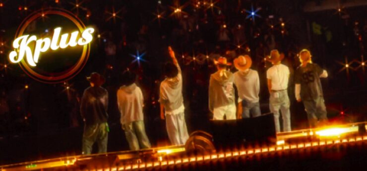 "Seven individuals stand on a stage facing an audience, with one raising a hand. They are wearing casual clothes and hats. Bright lights create a starry effect. The word 'Kplus' is visible in the upper left corner."