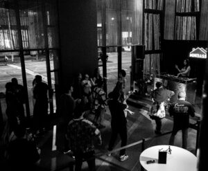 A lively indoor party scene with a DJ booth in the foreground featuring DJ equipment. A crowd of people is dancing energetically. The room is dimly lit with colorful lights creating a vibrant atmosphere. Large windows show a glimpse of the street outside.