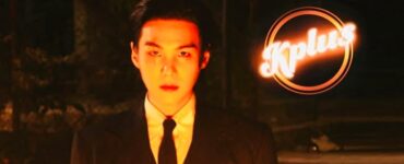 The image shows a person in a suit and tie standing in low light. The backdrop is dark, and the person is looking directly forward. The logo "Kplus" is brightly illuminated in the upper right corner.