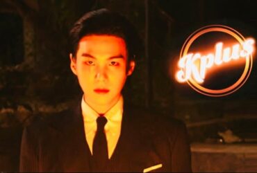 The image shows a person in a suit and tie standing in low light. The backdrop is dark, and the person is looking directly forward. The logo "Kplus" is brightly illuminated in the upper right corner.