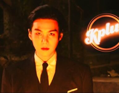 The image shows a person in a suit and tie standing in low light. The backdrop is dark, and the person is looking directly forward. The logo "Kplus" is brightly illuminated in the upper right corner.