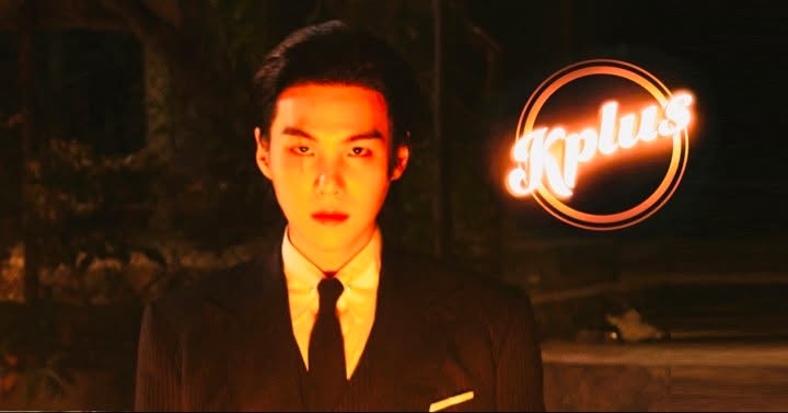 The image shows a person in a suit and tie standing in low light. The backdrop is dark, and the person is looking directly forward. The logo "Kplus" is brightly illuminated in the upper right corner.