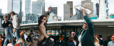 A crowded ferry deck with people taking photos and mingling, set against the backdrop of Seattle's skyline featuring tall buildings and landmarks. The top of the ferry's structure is visible, flying flags.