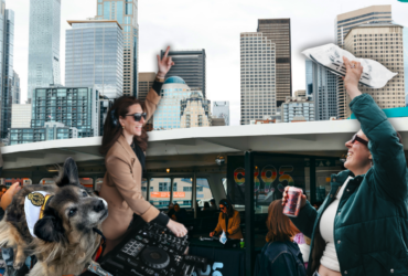 A crowded ferry deck with people taking photos and mingling, set against the backdrop of Seattle's skyline featuring tall buildings and landmarks. The top of the ferry's structure is visible, flying flags.