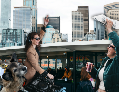 A crowded ferry deck with people taking photos and mingling, set against the backdrop of Seattle's skyline featuring tall buildings and landmarks. The top of the ferry's structure is visible, flying flags.